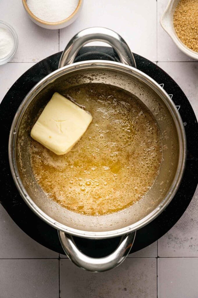 A stick of butter melting in a saucepan on a stovetop, with sugar and other ingredients in bowls nearby—everything you need to start making delicious Butter Pecan Cookies.