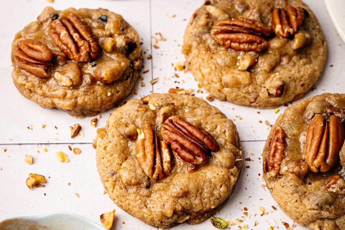 Four Butter Pecan Cookies with whole pecan halves on top are arranged on a light-colored surface, with small crumbs scattered around them.