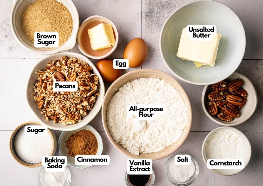 Bowls filled with baking ingredients for Butter Pecan Cookies, including brown sugar, sugar, flour, pecans, butter, eggs, cinnamon, vanilla extract, baking soda, salt, and cornstarch arranged on a counter.