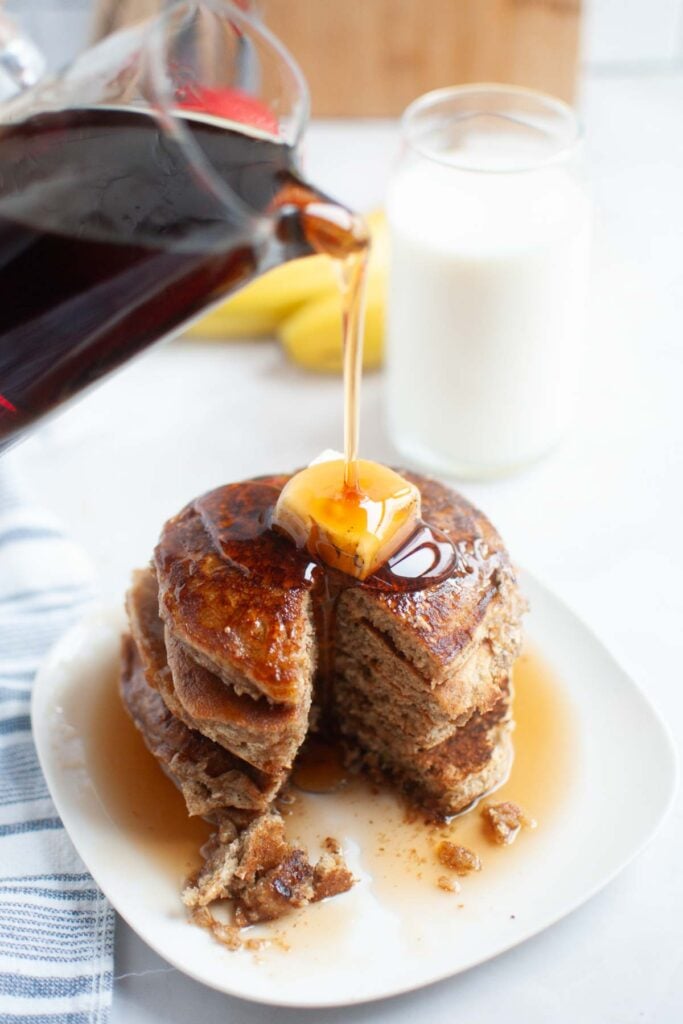 A stack of buttermilk oatmeal pancakes topped with butter and syrup, with syrup being poured from a pitcher. A glass of milk and sliced banana are in the background.