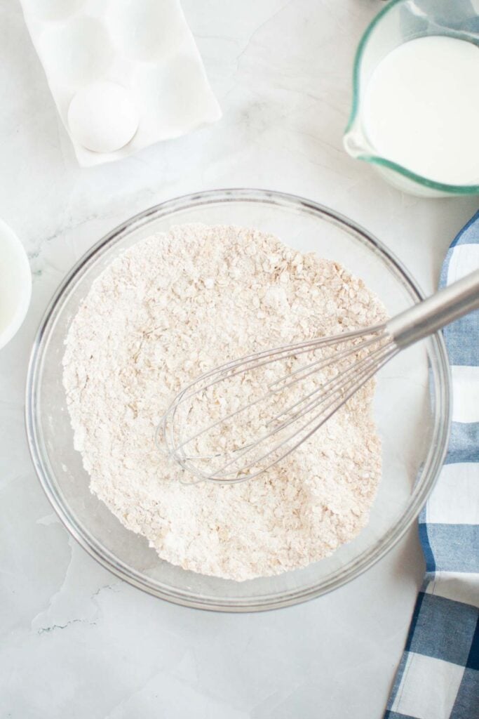 A glass bowl with flour mixture and a whisk on a countertop, ready to make buttermilk oatmeal pancakes, sits next to a cup of milk, an egg carton, and a blue checkered cloth.
