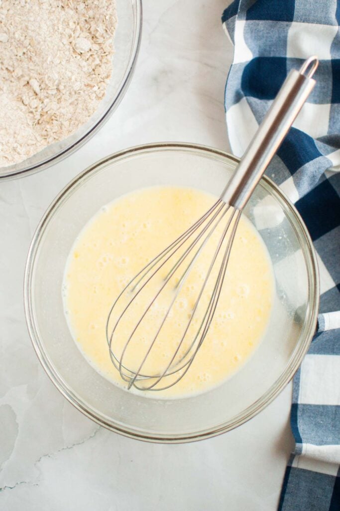 A glass bowl with beaten eggs and a whisk, next to a bowl of oats—perfect for making buttermilk oatmeal pancakes—and a blue-and-white checkered cloth on a marble surface.