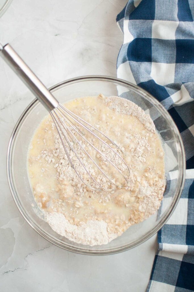 A glass bowl with flour, oats, and milk being mixed with a metal whisk—perfect for making buttermilk oatmeal pancakes—sits next to a blue and white checkered cloth on a light surface.