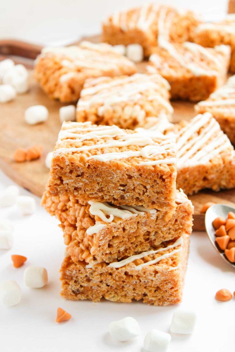 Three stacked Butterscotch Rice Treats drizzled with white icing, surrounded by mini marshmallows and butterscotch chips, with more treats on a wooden board in the background.