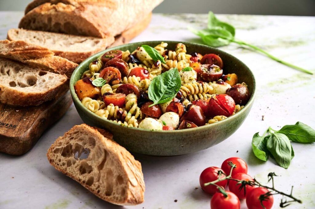 A bowl of Caprese Pasta Salad with cherry tomatoes, mozzarella, basil, and dressing is placed next to slices of crusty bread and fresh basil leaves on a white surface.
