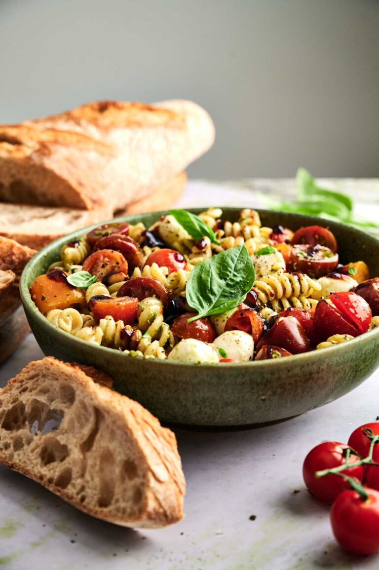 A bowl of Caprese Pasta Salad with cherry tomatoes, mozzarella, and basil sits on a table next to sliced baguette and fresh tomatoes.