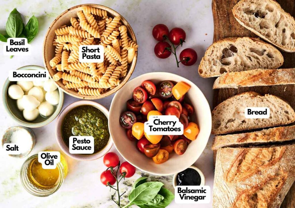 Various ingredients for Caprese Pasta Salad, including short pasta, cherry tomatoes, bocconcini, basil leaves, pesto sauce, olive oil, balsamic vinegar, salt, and bread are beautifully arranged on a white surface.