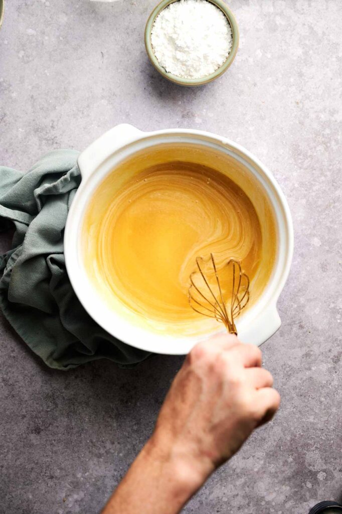 A hand whisks the chess pie filling until smooth and pale yellow, with powdered sugar waiting nearby.
