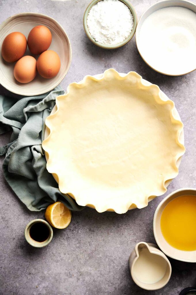An unbaked pie crust with crimped edges sits ready in a pan, surrounded by eggs, melted butter, sugar, and other chess pie ingredients.