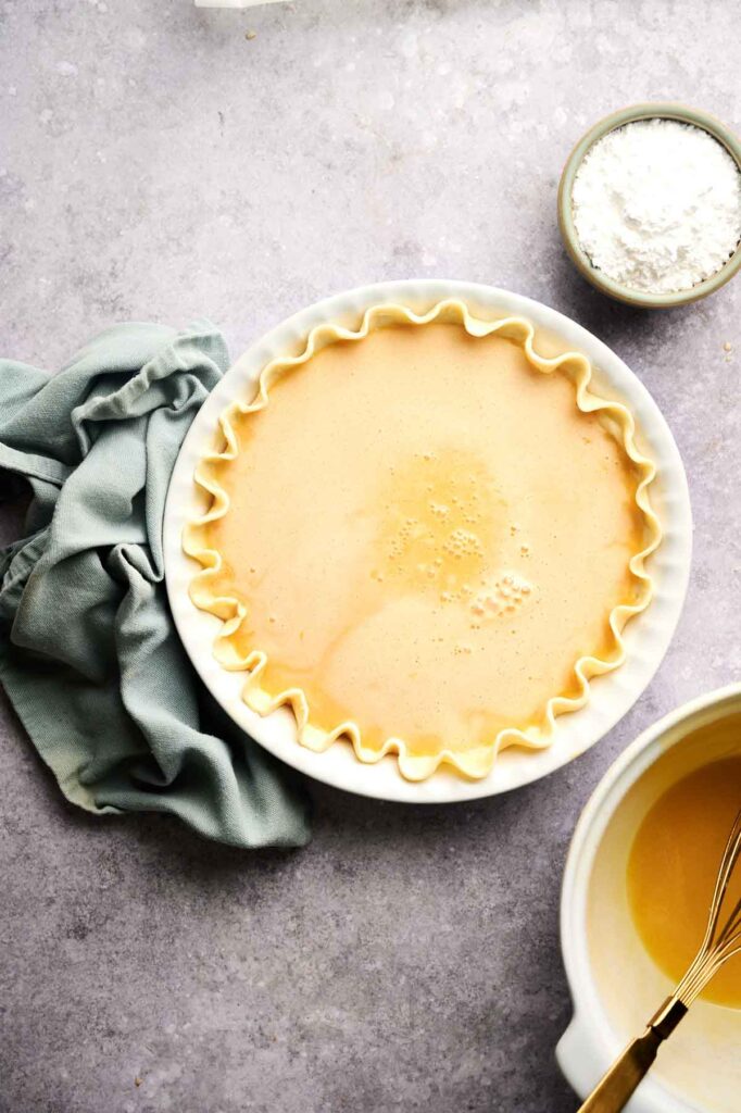 Unbaked Chess Pie with crimped edges in a white dish, a green cloth to the side, a bowl of powdered sugar, and a bowl with whisked filling and a whisk nearby.