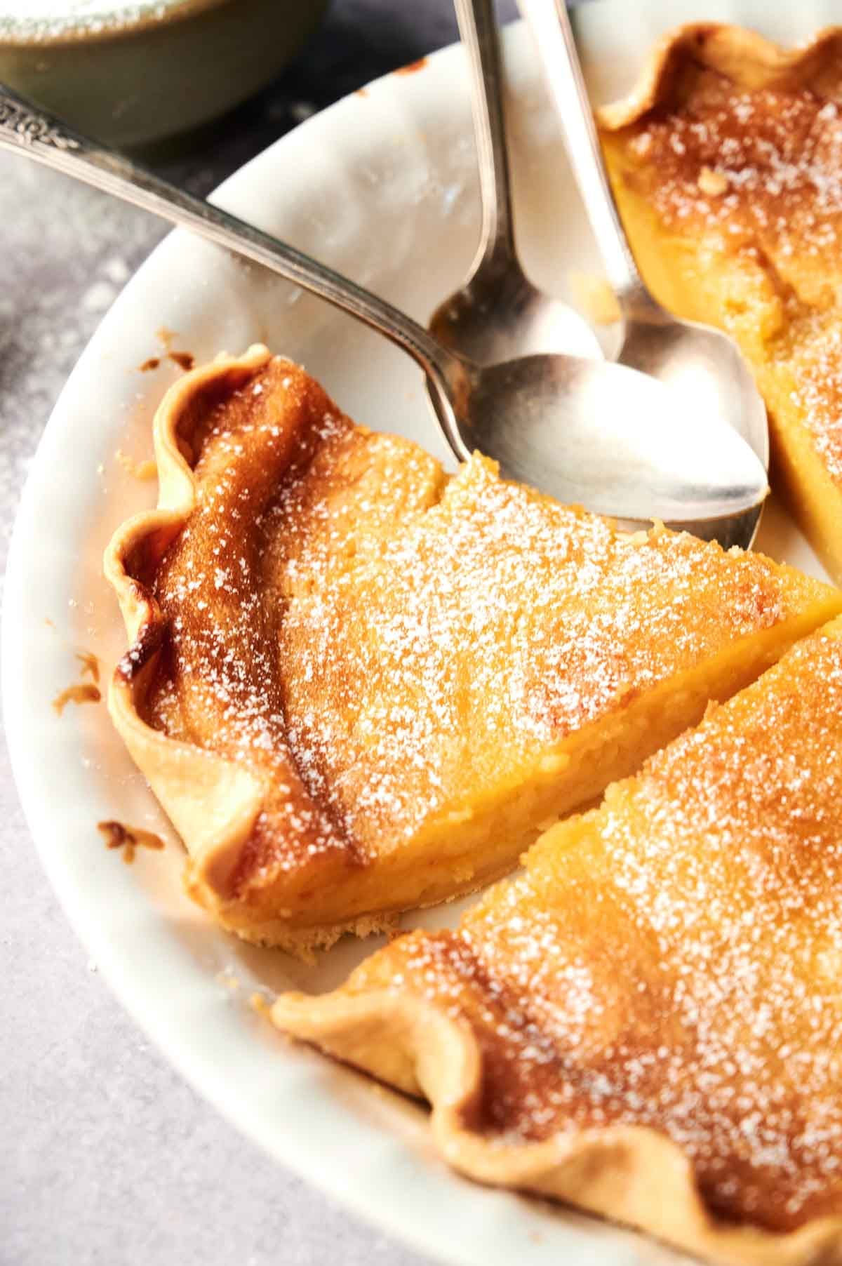 Close-up of sliced Chess Pie with a lightly browned crust, dusted with powdered sugar, and three metal spoons resting in the dish.