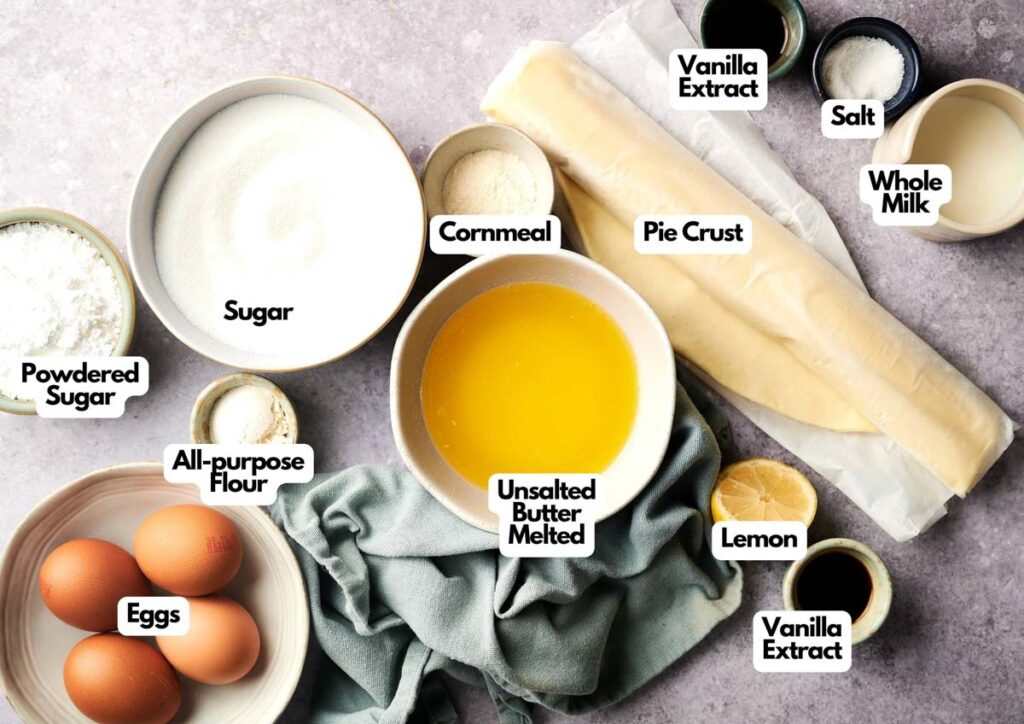Top-down view of labeled ingredients for baking Chess Pie, including pie crust, sugar, cornmeal, melted butter, eggs, flour, powdered sugar, vanilla extract, salt, whole milk, and lemon.
