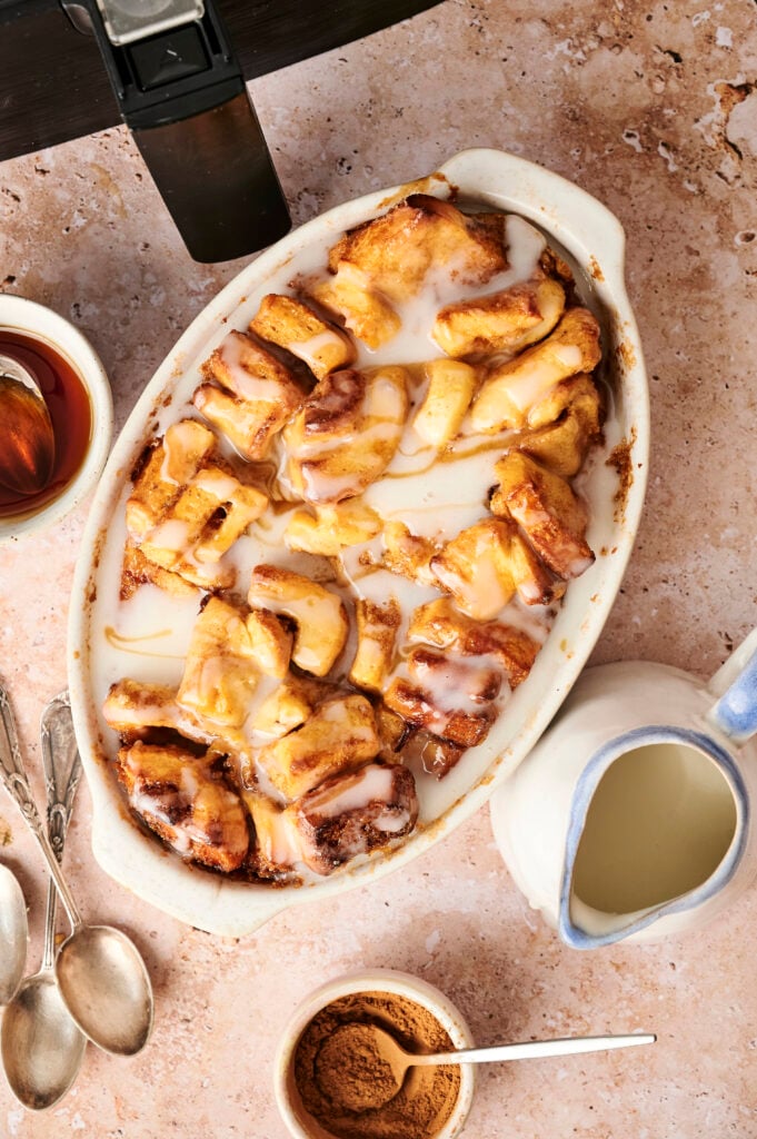 An irresistible Air Fryer Cinnamon Roll Casserole baked to perfection, featuring gooey cinnamon roll pieces topped with icing and surrounded by syrup, cream, cinnamon, and three spoons on a marble surface.