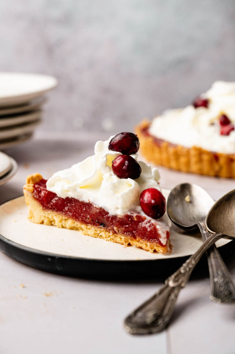 A slice of cranberry curd tart topped with whipped cream and whole cranberries sits on a plate beside two spoons, with more pie and stacked plates in the background.