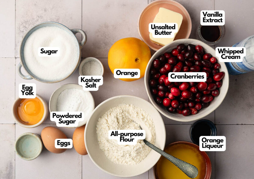 Top view of ingredients for a Cranberry Curd Tart on a countertop, labeled: sugar, kosher salt, egg yolk, powdered sugar, eggs, all-purpose flour, orange, unsalted butter, cranberries, vanilla extract, whipped cream, orange liqueur.