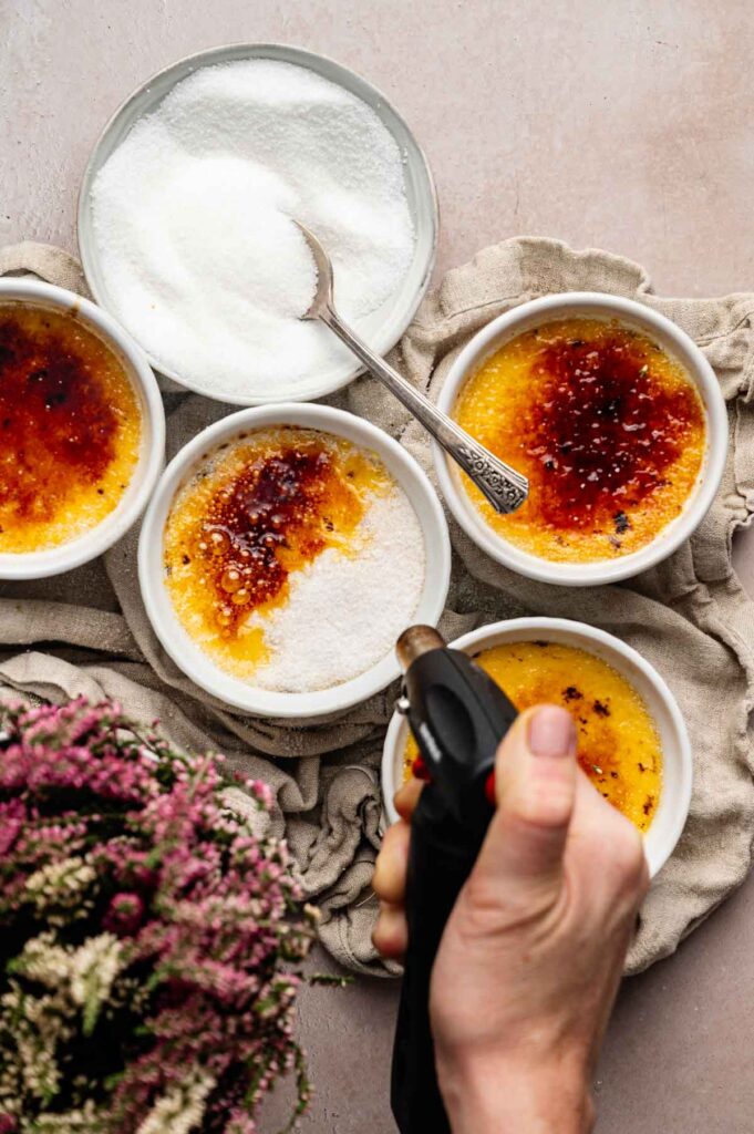 A hand uses a torch to caramelize sugar on top of classic cr&egrave;me br&ucirc;l&eacute;e in small white ramekins, with a bowl of sugar and spoon nearby.