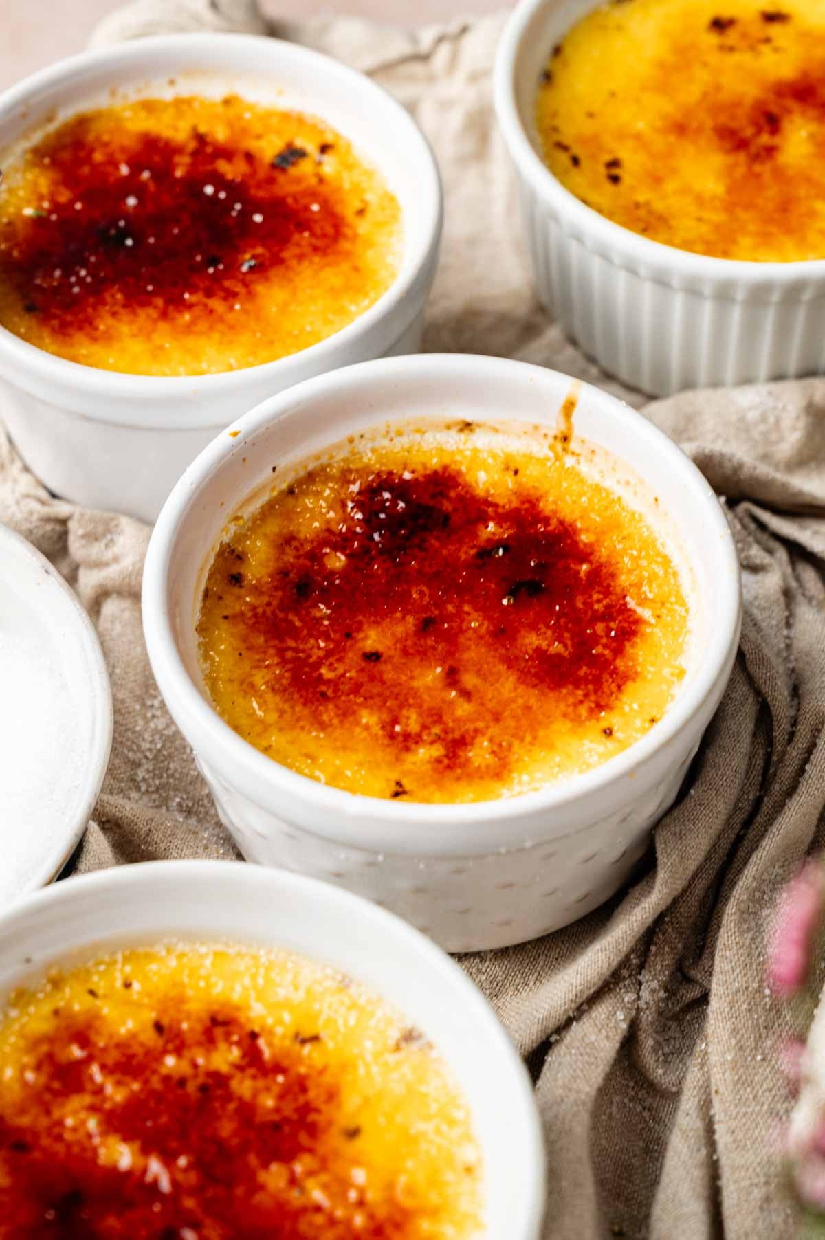 Four ramekins filled with rich Cr&egrave;me Br&ucirc;l&eacute;e, each topped with a caramelized sugar crust, are arranged on a beige cloth.