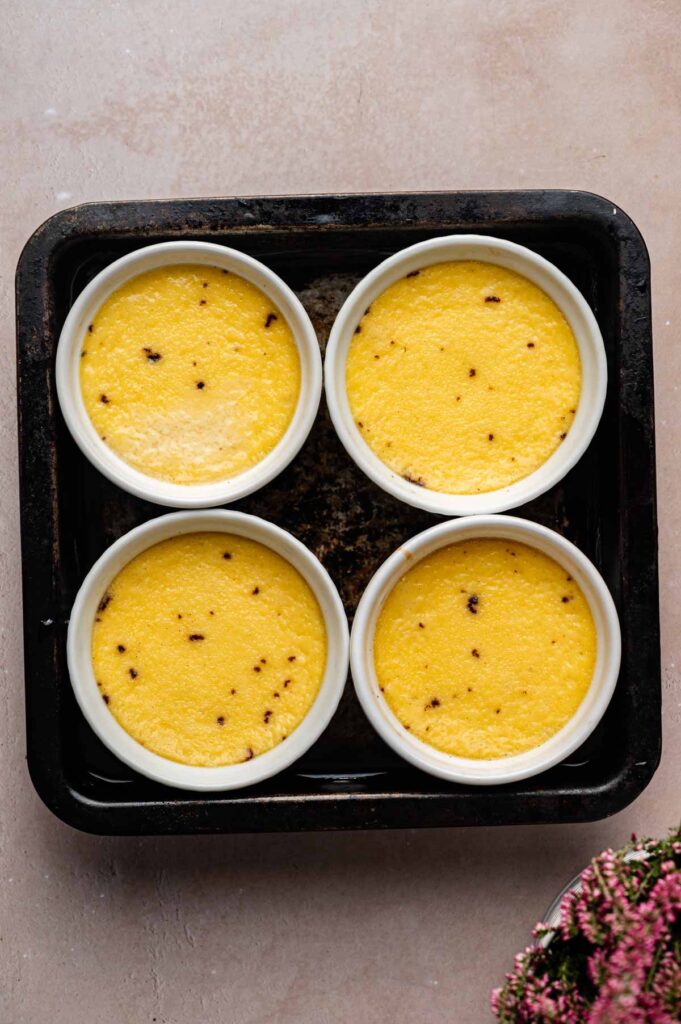 Four ramekins of Cr&egrave;me Br&ucirc;l&eacute;e, their yellow custard dotted with brown specks, are arranged in a black baking tray on a light surface.