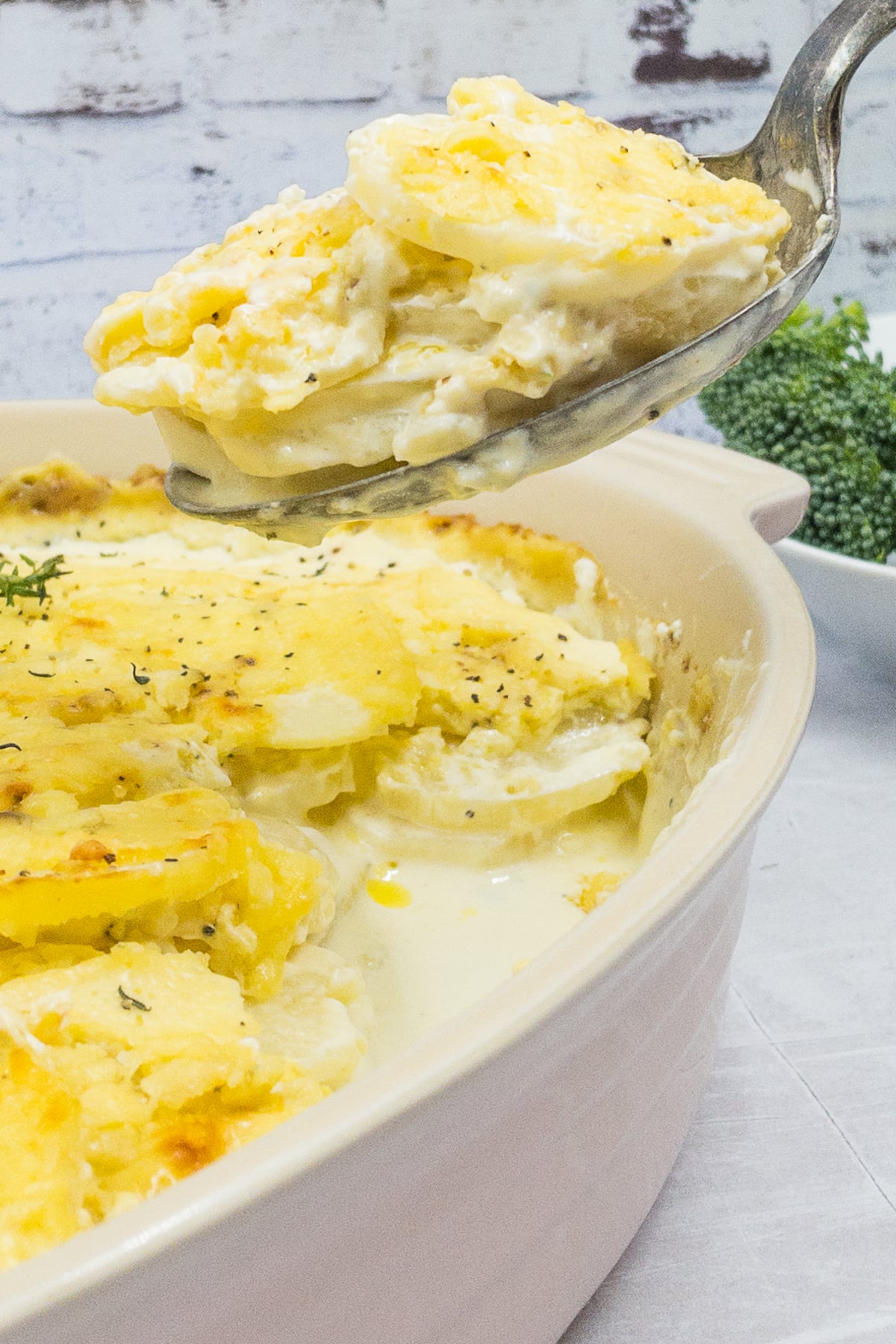 A spoon is lifting a serving of creamy Dauphinoise Potatoes from a baking dish, with a bowl of broccoli in the background.