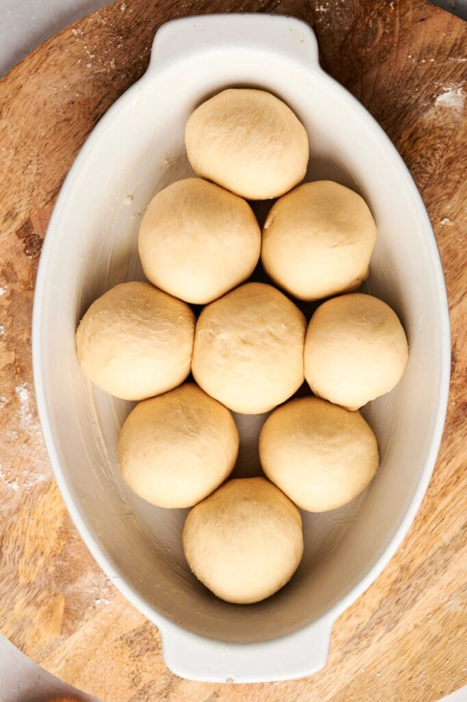 Nine round dough balls—soon to become fluffy Air Fryer Dinner Rolls—are arranged in a white oval baking dish on a wooden surface.