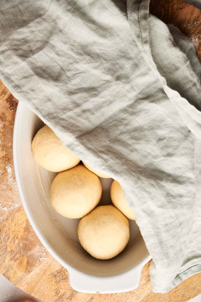 Four dough balls in a white dish are partially covered with a light gray cloth, resting on a wooden surface—perfect for transforming into soft Air Fryer Dinner Rolls.