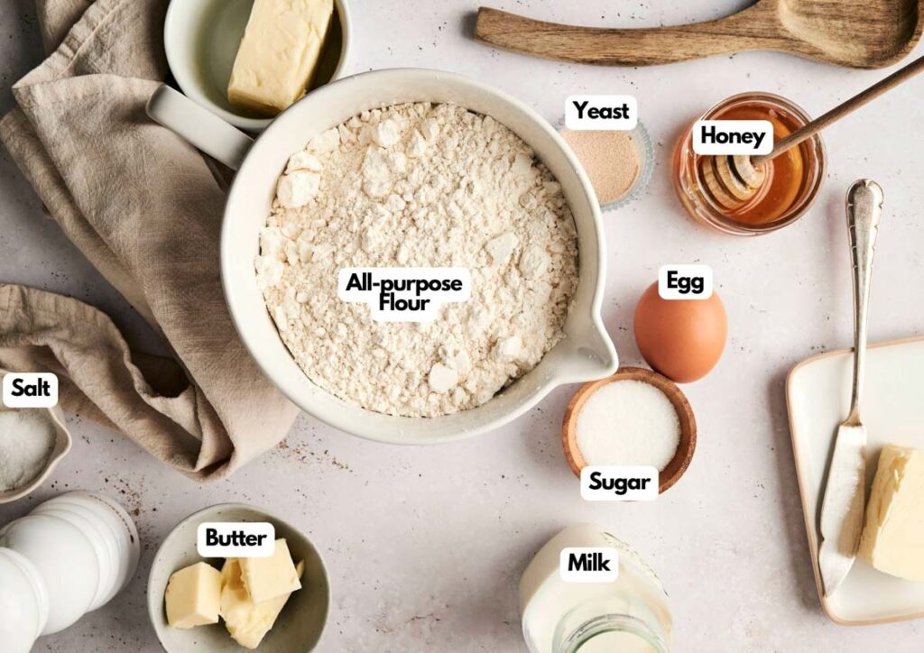 Top-down view of baking ingredients for Air Fryer Dinner Rolls on a countertop, including labeled all-purpose flour, yeast, honey, egg, sugar, milk, butter, and salt, with utensils and a cloth nearby.