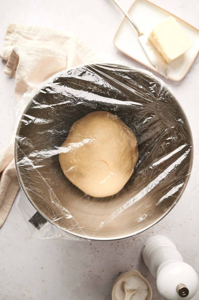 A ball of dough—soon to become fluffy Air Fryer Dinner Rolls—rests covered in plastic wrap in a mixing bowl on the countertop, with a butter dish, towel, and pepper grinder nearby.