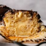 A close-up of a cut-open Boston Cream Pie Cupcake with chocolate icing and a creamy yellow filling, sitting on an unwrapped cupcake liner.