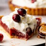 A slice of cranberry curd tart with whipped cream and whole cranberries on top, served on a plate with a spoonful of pie crust beside it.