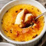 A close-up of classic Crème Brûlée in a white ramekin, its caramelized sugar topping cracked by a spoon breaking through the crispy surface.