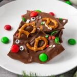 Reindeer Chocolate Bark pieces topped with pretzels, candy eyes, and festive sprinkles are served on a white plate, surrounded by red and green candies for a whimsical holiday treat.