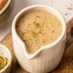 A small ceramic pitcher containing creamy brown vegetarian gravy, garnished with dried herbs, sits on a wooden board next to a bowl of seasoning.
