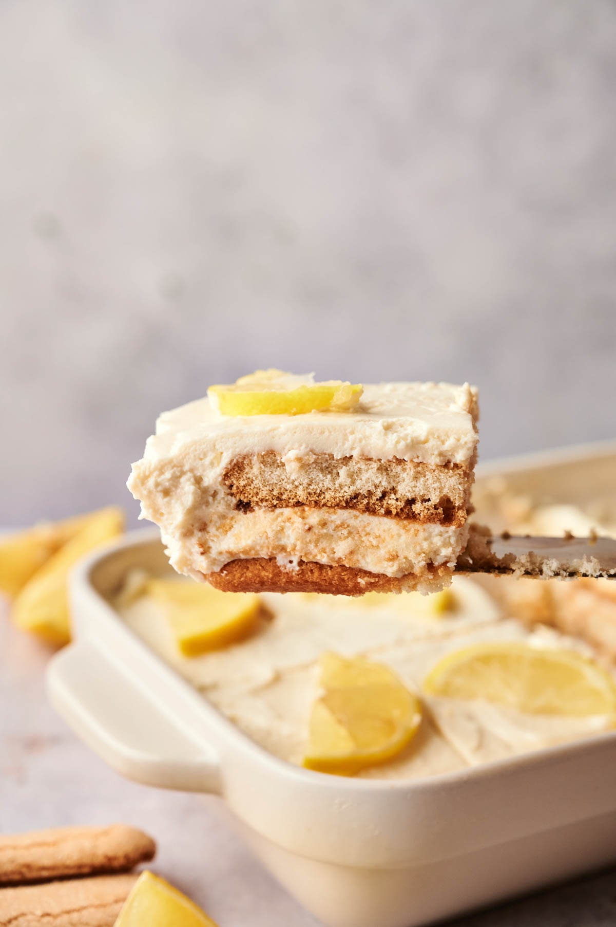 A spatula holds a square piece of Lemon Tiramisu, a layered lemon dessert with cream and cookies, above a baking dish filled with the same treat, garnished with fresh lemon slices.