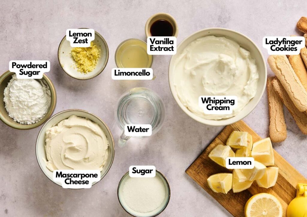 Ingredients for a Lemon Tiramisu dessert are arranged on a counter, including lemon zest, powdered sugar, mascarpone cheese, limoncello, water, sugar, vanilla, whipping cream, lemon, and ladyfinger cookies.