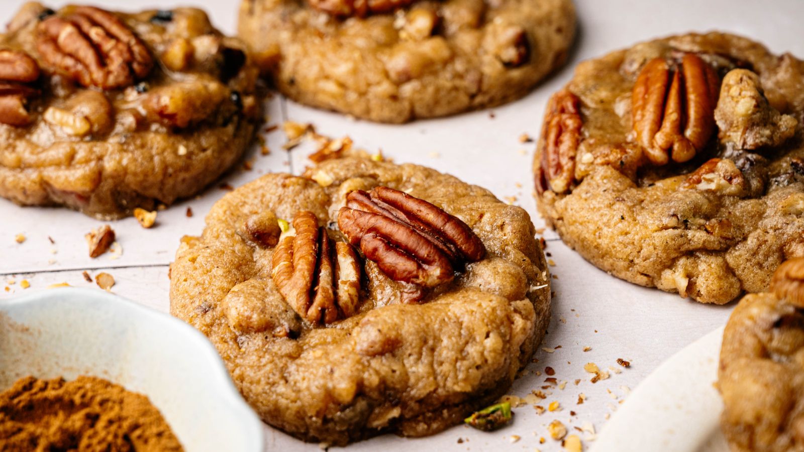Close-up of several pecan cookies on a light surface, each topped with a whole pecan. A small dish of cinnamon is visible in the corner.