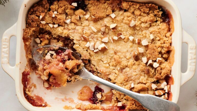 A baked fruit crumble topped with chopped nuts in a white dish, with a serving spoon and a portion missing.