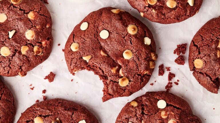 Several red velvet cookies with white chocolate chips on a sheet of parchment paper, one cookie has a bite taken out of it.