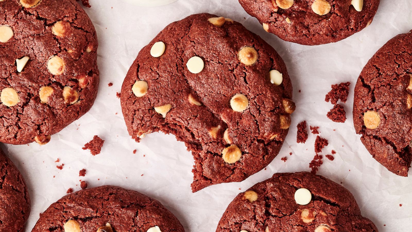 Several red velvet cookies with white chocolate chips on parchment paper, with one cookie in the center that has a bite taken out of it.