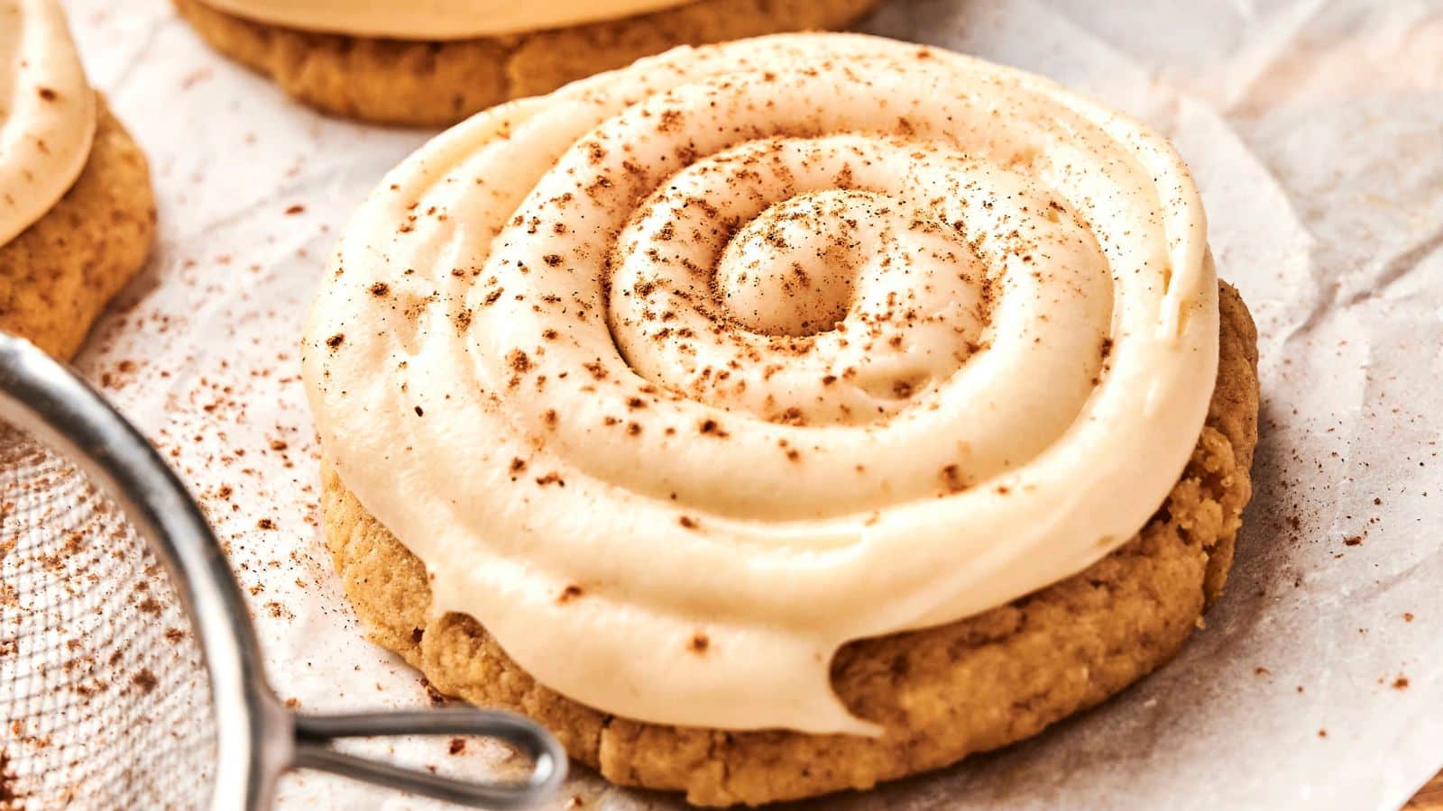 A close-up of a frosted cookie with a spiral swirl of icing and a dusting of cinnamon on top, placed on parchment paper.