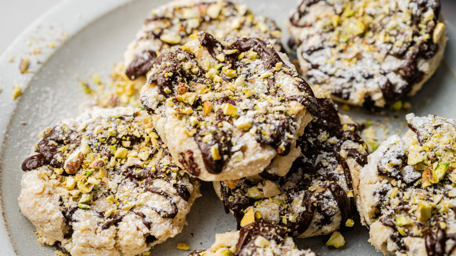 A plate of cookies topped with chopped pistachios, drizzled with chocolate, and dusted with powdered sugar.
