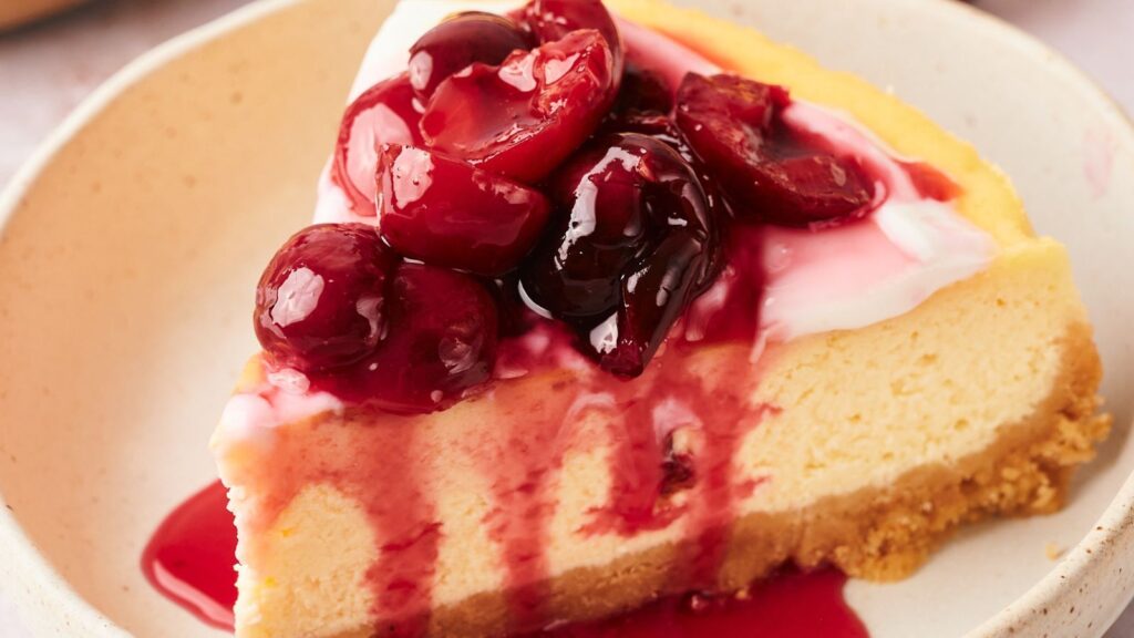 A slice of cheesecake with a graham cracker crust, topped with cherry sauce and whole cherries, on a beige plate.