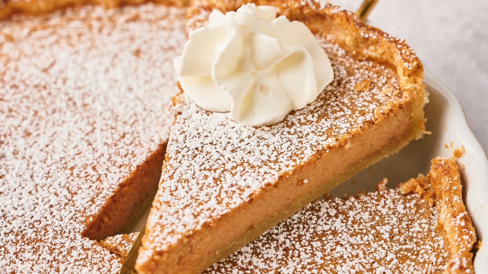 A slice of powdered sugar-dusted pie topped with a swirl of whipped cream is being lifted from a pie dish.