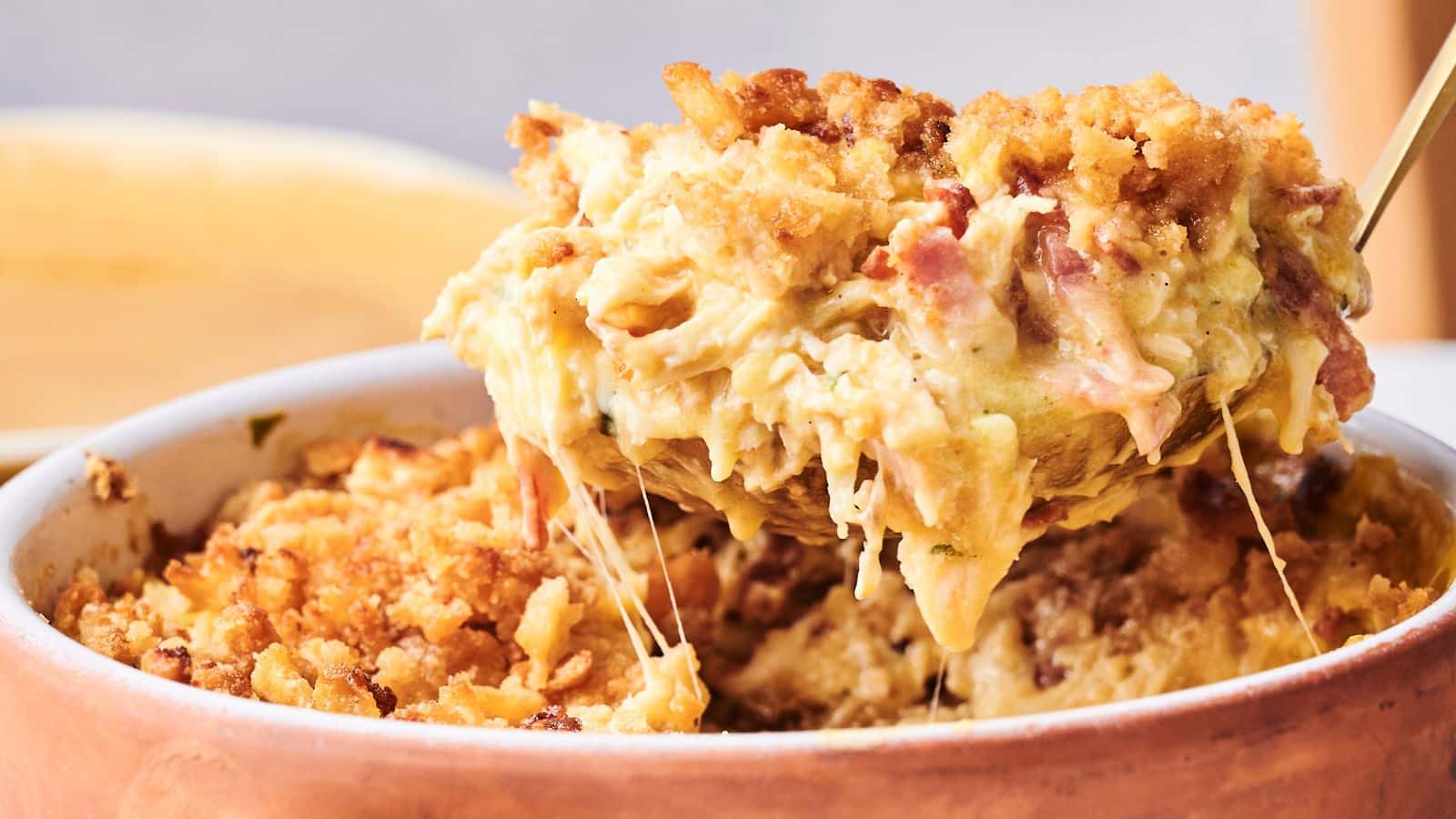 A spoon lifts a serving of baked macaroni and cheese with a golden breadcrumb topping and melted cheese from a casserole dish.