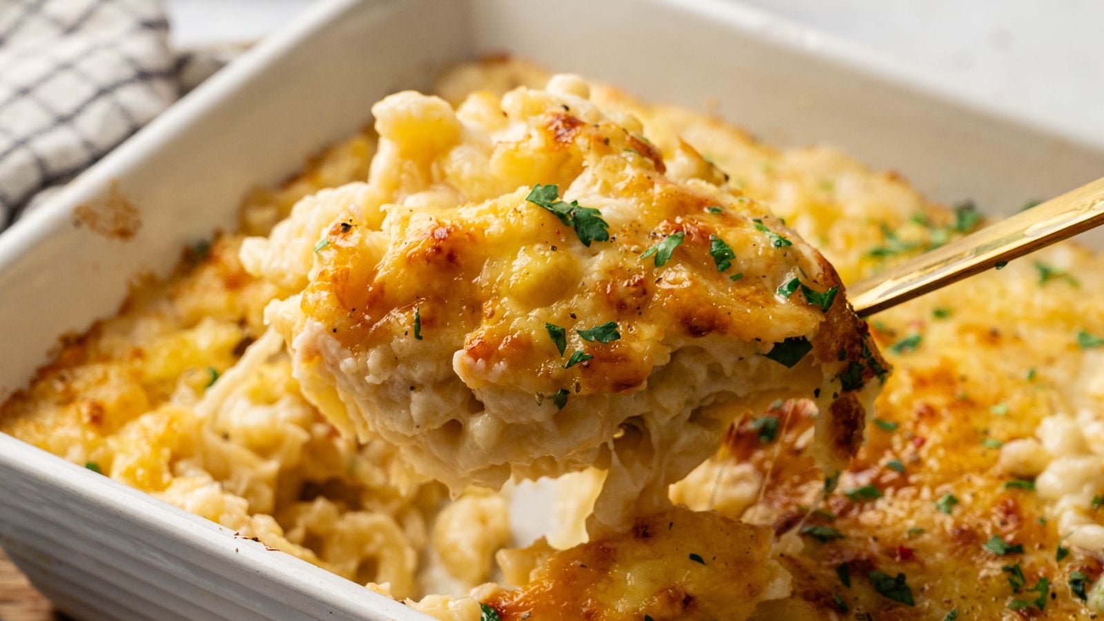 A close-up of a serving of baked macaroni and cheese being lifted from a casserole dish, topped with melted cheese and garnished with chopped herbs.