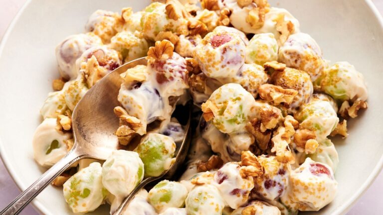 A bowl of grape salad with green and red grapes coated in creamy dressing, topped with chopped walnuts and brown sugar, with a spoon and fork.