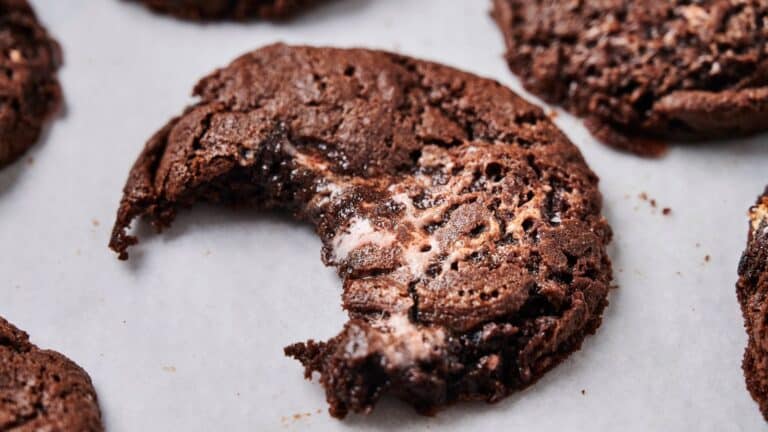 A close-up of a chocolate cookie with a bite taken out, showing a gooey marshmallow center, on a sheet of parchment paper.