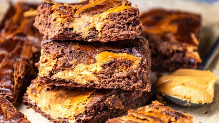 A stack of peanut butter swirl brownies with a spoonful of peanut butter beside them on a parchment surface.