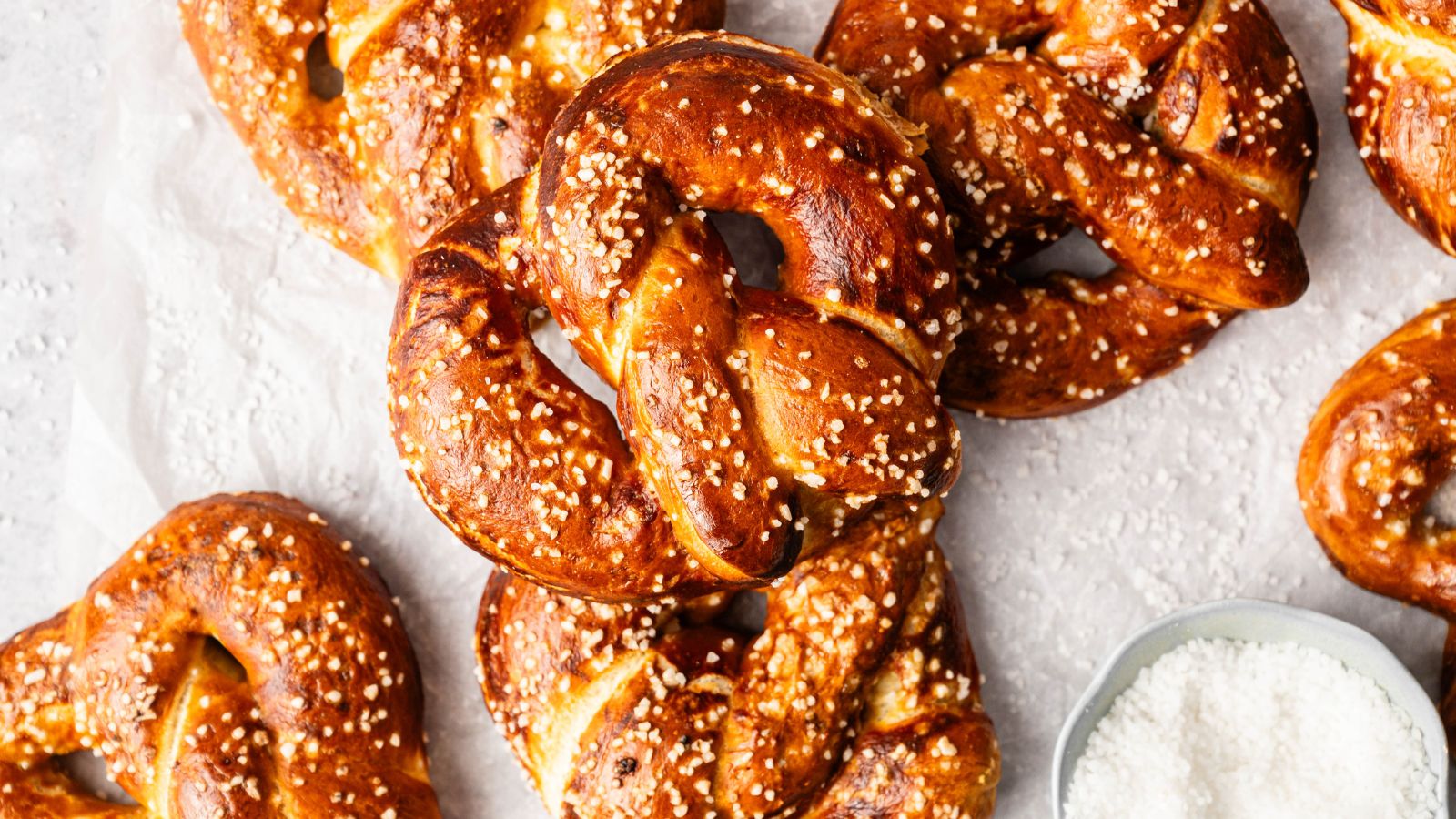 Several golden-brown pretzels with coarse salt are arranged on a light surface next to a small bowl of salt.