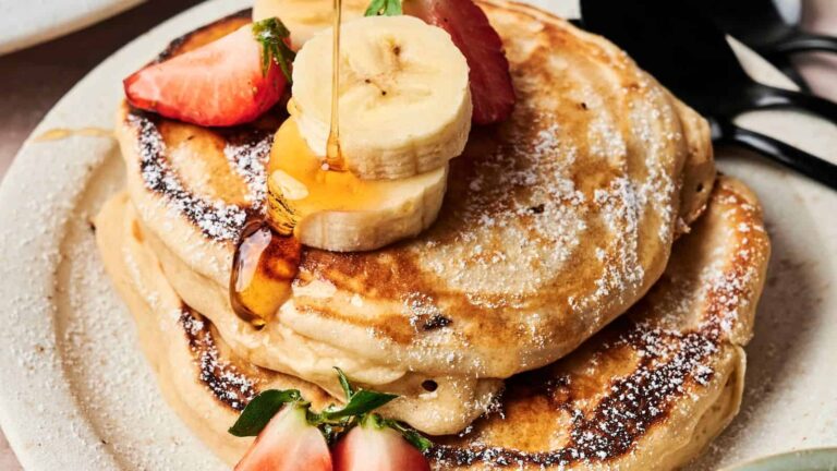 A stack of pancakes topped with sliced bananas, strawberries, and powdered sugar, with syrup being poured over them on a white plate.