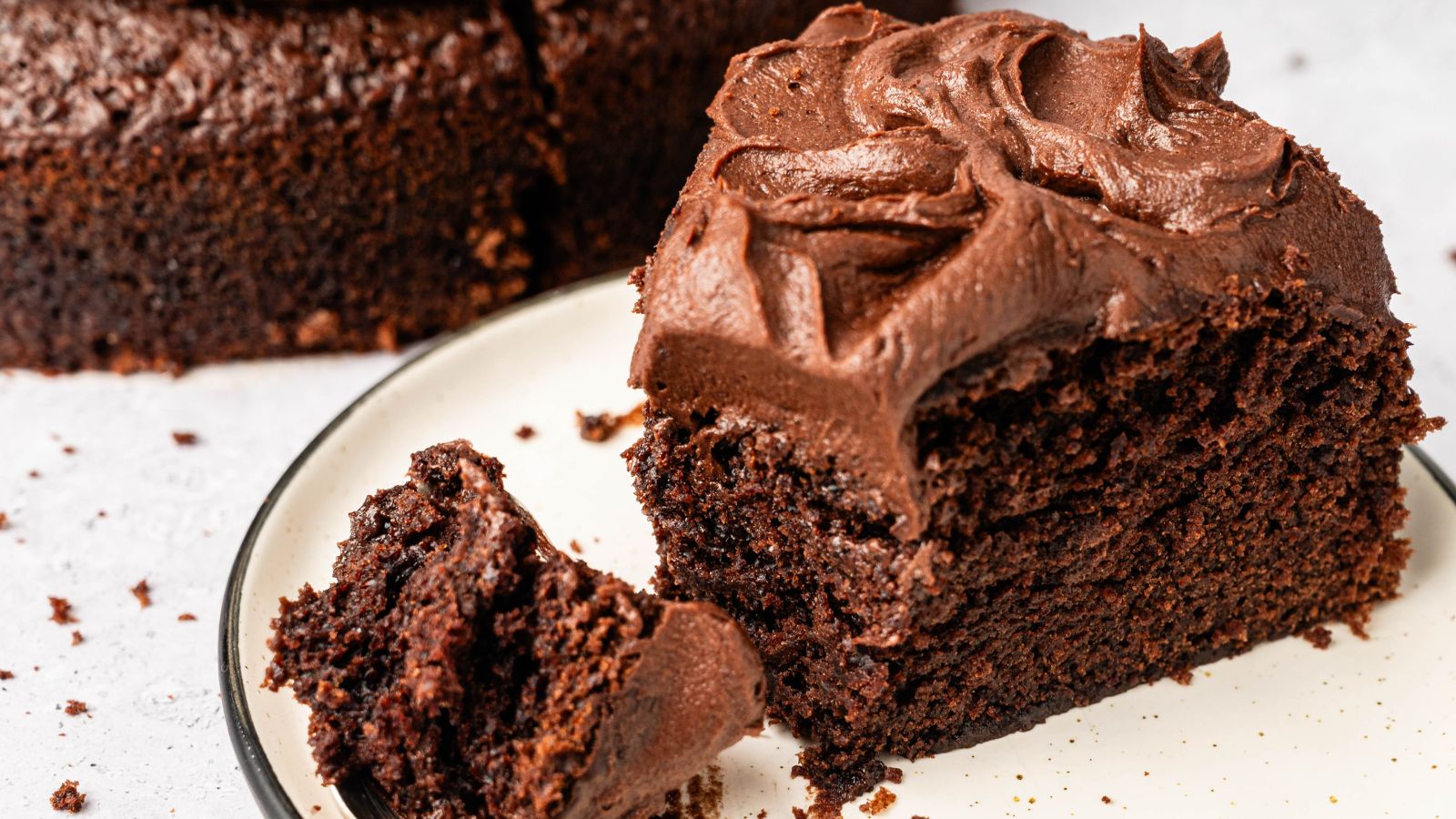 A slice of chocolate cake with chocolate frosting on a plate, with a small piece broken off and crumbs scattered around.